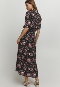 Robe maxi en tissu noir avec un motif floral en rose et blanc, manches courtes et détail de taille nouée. Dos fermé.
