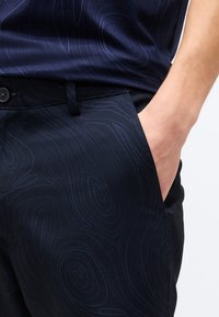 Χέρι ατόμου στην τσέπη σκούρου ναυτικού παντελονιού με διακριτικό τοπογραφικό μοτίβο, συνδυασμένο με αντίστοιχο πουκάμισο σε σκούρο ναυτικό χρώμα.