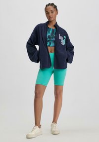 Marineblaues Fleece-Jackett mit Reißverschluss und einem lila Akzent-Logo, kombiniert mit einem türkisfarbenen Crop-Top und passenden Radshorts. Weiße Sneakers runden den Look ab.