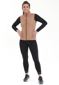 Gepolsterte, ärmellose, hellbraune Weste mit hohem Kragen, kombiniert mit einem langärmeligen schwarzen Oberteil und schwarzen Leggings, die kontrastierende Akzente aufweisen.
