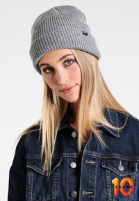 Szary dzianinowy beanie z prążkowaną teksturą, z małą czarną metką logo. Noszony przez osobę z długimi, prostymi włosami na jednolitym tle.