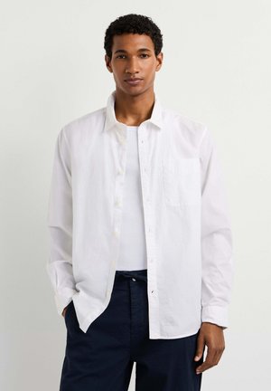 Junger Mann mit kurzen lockigen Haaren, der ein weißes Hemd mit Knöpfen über einem weißen Tanktop und dunklen Hosen trägt, stehend vor einem einfarbigen hellen Hintergrund.
