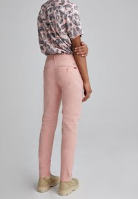 Light pink fitted pants with back pockets and subtle branding, paired with a short-sleeve patterned shirt. Shoes are beige.