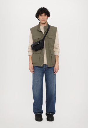 Vesta verde măsliniu peste un pulover bej, asociat cu blugi evazați de culoare albastru deschis și pantofi slip-on negri, având o geantă crossbody neagră.