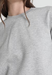 Sudadera gris hecha de un tejido suave, que presenta un cuello redondo acanalado y un diseño de hombros sin costuras. Textura lisa con un detalle mínimo.