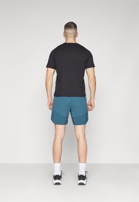 Svart kortärmad skjorta, teal shorts med ett strukturerat mönster, vita strumpor och svarta sneakers. Vyn visar baksidan av outfiten.
