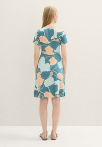 Frau steht mit dem Rücken zur Kamera und trägt ein knielanges Kleid mit einem abstrakten floralen Muster in Blau, Pfirsich und Weiß sowie Sandalen vor einem schlichten Hintergrund.