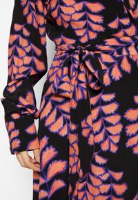 Robe à manches longues avec base noire et motif de feuilles orange vif souligné de violet, dotée d'une ceinture nouée à la taille et de manches retroussées.