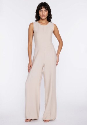 Kobieta ubrana w bezrękawnikowy beżowy kombinezon z szerokimi nogawkami, stojąca na tle jednolitego białego tła, z jedną ręką na biodrze.