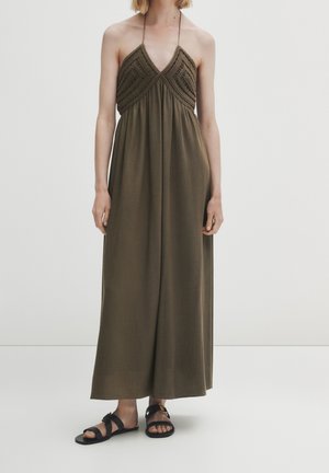 Kobieta ubrana w oliwkowozieloną sukienkę maxi na szyję z szydełkowanym gorsetem i czarne płaskie sandały, stojąca na tle jednolitej białej ściany.