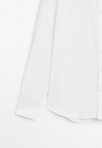 Hvid langærmet skjorte med knaplukning fortil, spidse kraver og rundet kant. Stoffet synes glat med en let glans.
