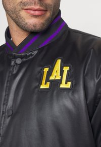 Chaqueta de satén negra con cuello acanalado a rayas púrpura y negra, bordado "LAL" en amarillo, y cierre de botón a presión.