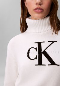 Woman wearing white ribbed turtleneck sweater with large black "cK" logo on front, brown hair partially visible, gray background.