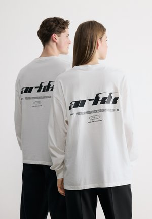 Due persone che indossano maglie a manica lunga bianche con la scritta nera in grassetto "arikk" e un piccolo logo stampato sul retro, su uno sfondo uniforme.