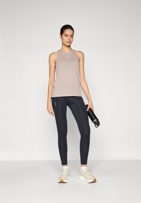 Débardeur taupe clair avec un col rond, associé à un legging noir taille haute et des chaussures de sport beiges. Une bouteille d'eau est tenue.