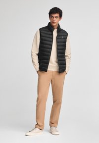 Svart vadderad väst med hög krage över en beige sweatshirt, kombinerad med ljusbruna byxor och krämfärgade sneakers. Enkel design, inga synliga knäppningar.