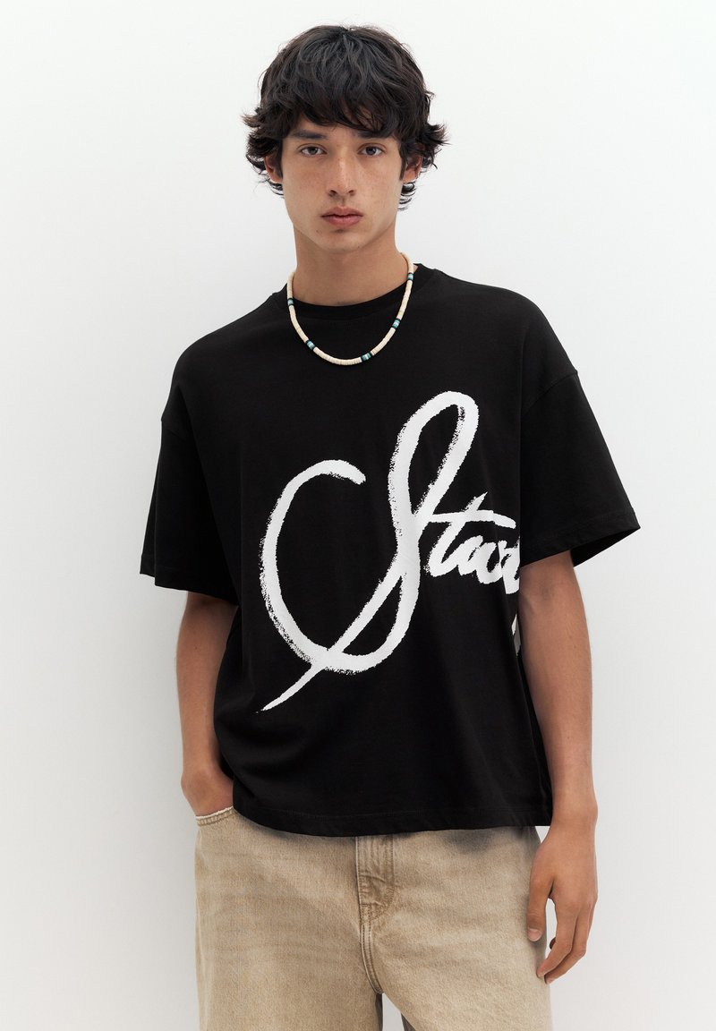 Jeune homme aux cheveux foncés portant un t-shirt noir oversize avec un grand texte blanc en cursive et un pantalon beige clair, debout sur un fond blanc.