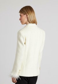 Mujer con cabello castaño claro que lleva un blazer blanco de pelaje suave con botones de perla y puños con plumas, de perfil frente a un fondo sencillo.