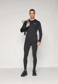 Giovane uomo che indossa abbigliamento atletico nero e sneakers, con uno zaino beige su una spalla, in piedi contro uno sfondo chiaro e uniforme.