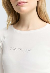 Weißes Baumwollhemd mit langen Ärmeln und Rundhalsausschnitt, das ein silbernes "TOM TAILOR"-Logo in gepunktetem Design über der Brust zeigt.