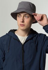Grå bucket hat lavet af stof, med en bred skygge og blød tekstur, parret med en marineblå jakke med lynlås foran og snoretræk.