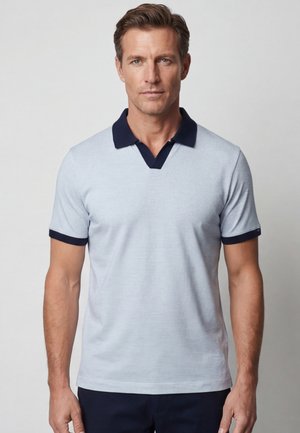 Man met kort bruin haar draagt een lichtblauw poloshirt met korte mouwen en een donkerblauwe kraag en manchetten, staand tegen een effen grijze achtergrond.