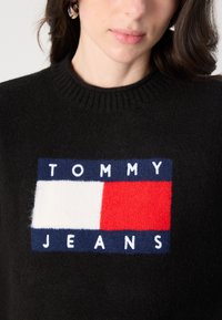 Svart strikket genser med et rektangulært logo som har blå, hvite og røde seksjoner og teksten "TOMMY JEANS" i hvite bokstaver.