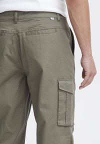 Pantaloni de tip cargo de culoare verde măsliniu, cu o croială relaxată, având buzunare laterale, un buzunar tip patch pe picior și o etichetă cusută pe talie.