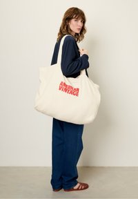 Sac fourre-tout beige au toucher doux, avec un logo rouge "AMERICAN VINTAGE" bien en vue. Grande forme décontractée avec de larges anses.