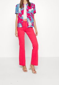 Kortärmad blå skjorta med stort rosa och vitt blommönster, kombinerad med klarröda utsvängda byxor och vita remmodeller med hög klack.