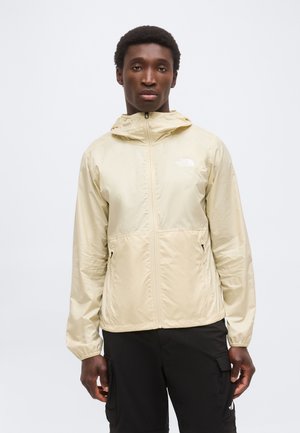 Mann trägt beige Kapuzenjacke mit Reißverschluss und schwarze Cargohosen, steht vor einfarbigem hellgrauem Hintergrund.