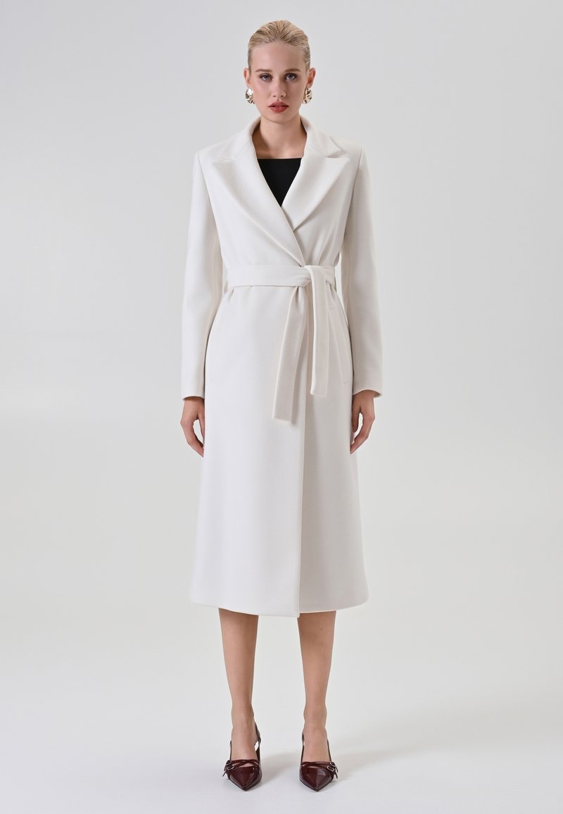 Donna che indossa un lungo cappotto bianco con cintura sopra un top nero, con tacchi a punta bordeaux, in piedi contro uno sfondo chiaro e uniforme.
