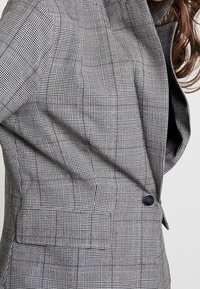 Blazer a scacchi grigi con una trama testurizzata, dotato di colletto a punte, chiusura con un solo bottone e tasca laterale.