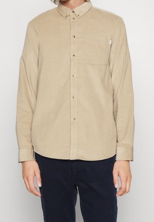 Beige kordfløjls-skjorte med knapper, krave og brystlomme, båret med mørke kordfløjlsbukser på en person fra nakke til hofter.