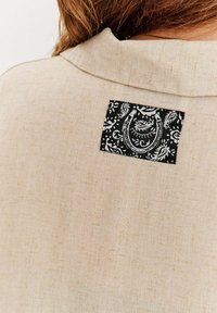 Béžová lněná košile s černým vzorovaným záplatou na zádech s bílými květinovými motivy. Texturovaná tkanina, jednoduchý design límce.