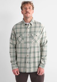 Chemise en flanelle à carreaux verts et crème avec des manches longues, un devant boutonné et deux poches poitrine. Présente un tissu texturé et chaud.