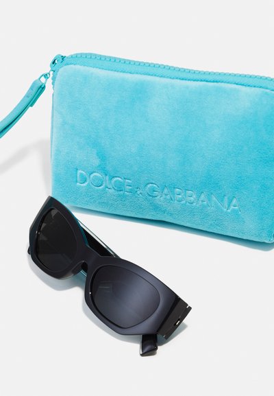 Occhiali da sole neri e oversize con finitura opaca, esposti accanto a un caso in velluto turchese con logo del marchio impressa.