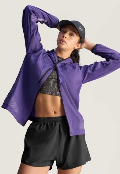 Chaqueta ligera morada con cremallera, paneles de malla y capucha, combinada con pantalones cortos negros de cintura elástica. Top corto texturizado debajo.