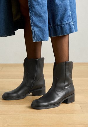 Bottes en cuir noires mi-mollet avec fermetures éclair sur les côtés, portées avec des collants noirs transparents et une jupe en jean bleu sur un sol en bois.