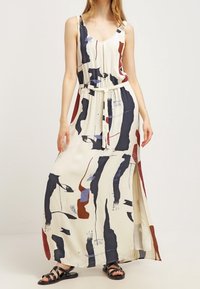 Robe maxi crème avec des motifs abstraits bleu marine, rouge et bleu. Elle dispose de bretelles spaghetti, d'une taille à cordon de serrage et d fentes sur les côtés.