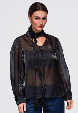 Femme portant un chemisier noir transparent à manches longues avec des détails floraux, associé à un jean bleu, souriante avec les cheveux attachés.