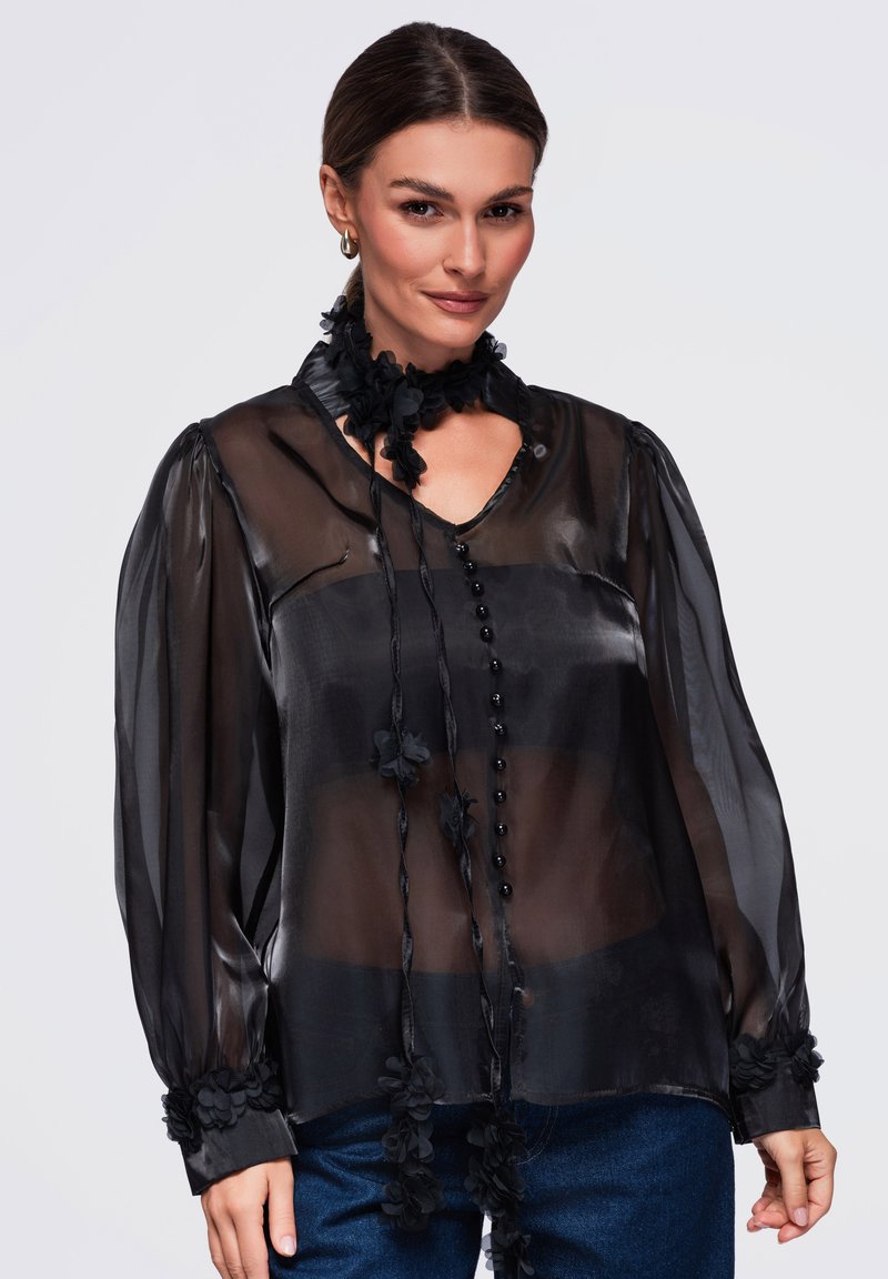 Femme portant un chemisier noir transparent à manches longues avec des détails floraux, associé à un jean bleu, souriante avec les cheveux attachés.