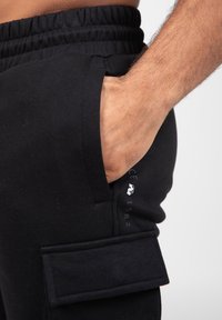 Black cotton sweatpants with an elastic waistband, side pocket, and a cargo pocket with flap. Features logo and year printed near the hem.