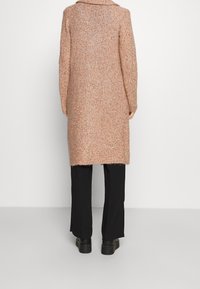 Larga chaqueta de mezcla de lana en rosa texturizado con motas de colores. Presenta un cuello ancho y un dobladillo deshilachado. Combinada con pantalones negros y botas gruesas.