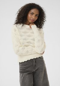 Pull en maille crème avec un motif de losanges ajouré, col rond côtelé et ourlet festonné, associé à un pantalon gris à coupe décontractée.
