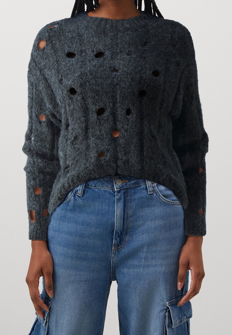 Personne portant un pull gris foncé en tissu duveteux avec plusieurs trous et un pantalon cargo en denim bleu clair avec des poches à l'avant.