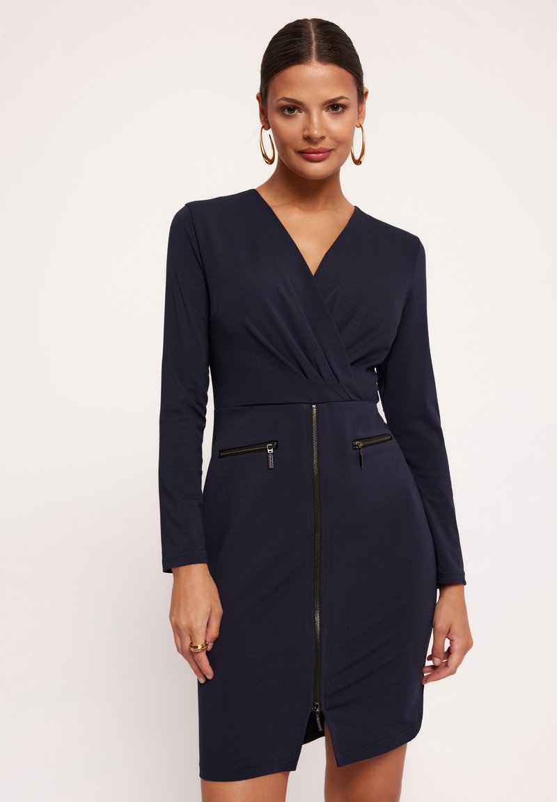 Granatowa sukienka kopertowa z długimi rękawami, z suwakiem z przodu, dwiema zapinanymi na zamek kieszeniami oraz dopasowanym krojem. Gładka tekstura materiału.
