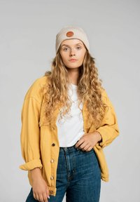 Żółta, oversize'owa kurtka, biała koszula, ciemnoniebieskie dżinsy oraz beżowa dziana czapka z okrągłym skórzanym detalem. Widoczne długie, kręcone włosy.