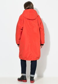 Parka rouge imperméable avec une capuche, un ourlet élastiqué et des manches longues. Associée à un jean foncé et des bottes noires, vue de dos.
