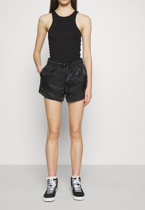 Femme debout les mains dans les poches, portant un débardeur noir sans manches, un short noir brillant, des chaussettes blanches et des baskets montantes noires.